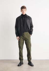 Casaco bombardeiro de couro, polo preto, calças cargo verde-oliva com bolsos laterais e sapatos pretos de atacadores. Design simples e funcional.