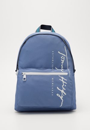 Niebieski plecak z białą kieszenią na zamek z przodu, białym logo i napisem Tommy Hilfiger oraz paskowanym uchwytem na górze.