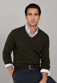 Mörkgrön v-ringad tröja i mjukt material, bärs över en ljusblå skjorta med krage. Modellen har ett svart bälte med en silverspänne.