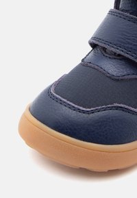 Sneaker bleu foncé avec une tige en cuir et textile, dotée de bandes velcro, d'accents cousus et d'une semelle en caoutchouc beige. Vue rapprochée de l'avant.
