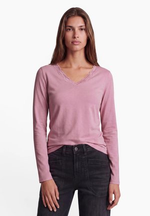 Jeune femme portant un haut rose à manches longues avec un col en V bordé de dentelle et un jean noir taille haute, debout devant un fond blanc.