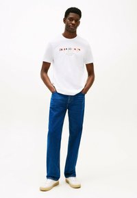 T-shirt bianco a maniche corte con grafica logo colorata, abbinato a jeans in denim blu e sneaker bianche. Design semplice e vestibilità rilassata.
