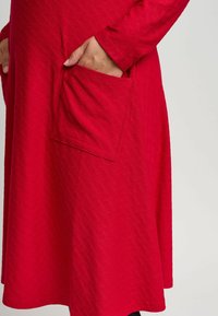 Röd klänning i texturerad tyg, med långa ärmar och en stor sidoficka. Designen har ett subtilt mönster genom hela plagget.