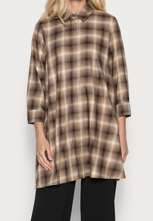 Kvinde iført brun og beige ternet skjorte med lange ærmer og løs pasform med krave, kombineret med sorte bukser, stående mod en ensfarvet baggrund.