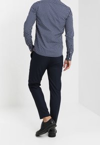 Man in een marineblauw overhemd met stippen en lange mouwen, donker slim-fit broek en zwarte nette schoenen die met zijn rug naar de camera staat.