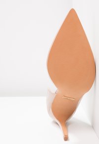 Escarpin à bout pointu en cuir verni nude avec un talon stiletto effilé et une semelle beige texturée, ornée d'un petit logo métallique.
