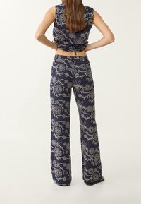 Vrouw met lang haar in een marineblauwe mouwloze top en wijde broek met witte bloemenborduursels, staand met handen op de heupen.