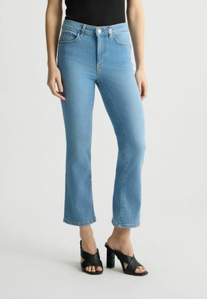 Helleblaue Jeans im Schlag mit hoher Taille, die das traditionelle Fünf-Taschen-Design und dezente Steppdetails aufweisen. Kombiniert mit schwarzen Absatz-Mules.