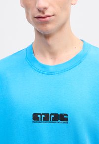 Tyrkysové bavlnené tričko s okrúhlym výstrihom. Na hrudi sa nachádza čierne logo "MADS NØRGAARD COPENHAGEN". Mäkká textúra.