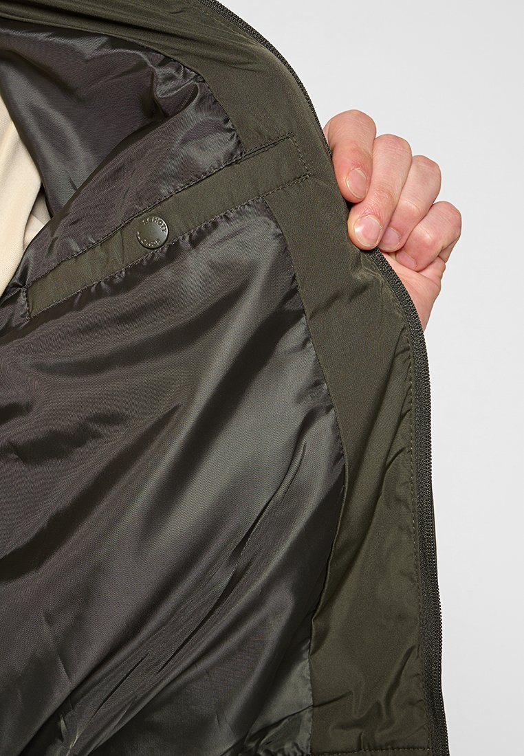 Intérieur de veste vert olive, avec une doublure lisse et brillante, fermeture éclair et un détail en métal circulaire sur la poche intérieure.