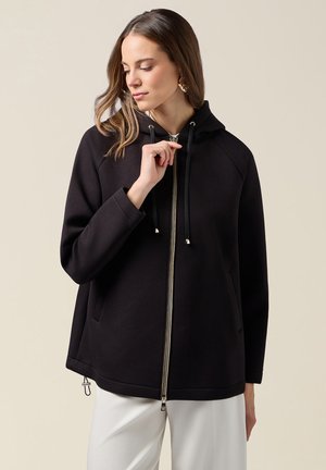 Vrouw draagt een zwart vest met ritssluiting en trekkoorden, samen met een witte broek, kijkt naar beneden en past de ritssluiting aan met haar rechterhand.