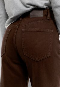 Közelkép egy személyről, aki barna farmernadrágot visel hátsó zsebekkel és derekán betűrt szürke pulóverrel.