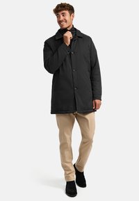 Abrigo acolchado negro con cierre de botones, cuello ancho y bolsillos laterales; llevado sobre pantalones beiges y botas negras. Textura de tela suave.