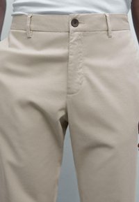 Beige pants with button closure and belt loops, worn with a tucked-in white shirt, shown from waist to mid-thigh.