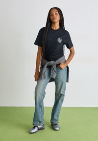 Black T-shirt with a round graphic. Paired with light blue jeans and gray sweatshirt tied at the waist. Silver sneakers complete the look.