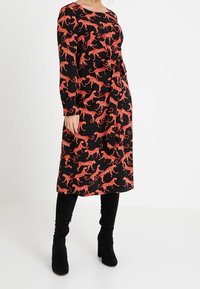 Schwarzes Kleid mit rotem Leopardenmuster, langen Ärmeln, rundem Ausschnitt und gebundenem Taillengürtel. Getragen mit knielangen schwarzen Stiefeln.