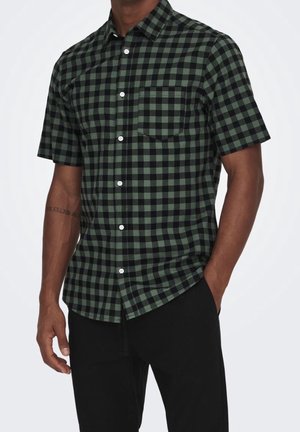 Homme portant une chemise à manches courtes à carreaux noir et vert foncé avec une poche à l'avant, associée à un pantalon noir, debout avec une main dans la poche.