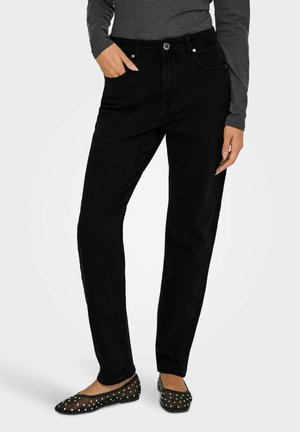 Person iført sorte high-waist jeans, grå langærmet top og sorte gennemsigtige flats med sølvnitter, står med den ene hånd i lommen.