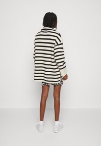 Personne aux cheveux tressés portant un pull oversize rayé noir et blanc, une jupe à motifs, des chaussettes blanches et des baskets blanches, debout, de dos.