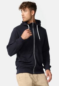 Marineblå hettegenser med glidelås, snor i hetten, frontlommer og ribbestrikkede mansjetter. Har en logo på brystet og sølvfarget glidelåsdetalj.