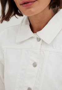 Femme portant une veste en denim blanche avec un col rabattu et des boutons-pression en métal, coupée sous le nez et au-dessus de la poitrine.