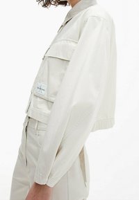 Zkrácená světle béžová bunda z měkkého materiálu, s límcem, dvěma předními kapsami a elastickými manžetami. Jemný detail s logem.