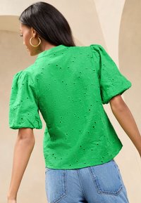 Donna che indossa una blusa verde ricamata con maniche a sbuffo e jeans azzurri, in piedi di spalle alla fotocamera.