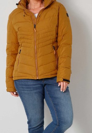 Modèle portant une veste à capuche matelassée jaune moutarde avec des poches zippées et un jean bleu.