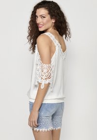 Blusa bianca con maniche a freddo bordate in pizzo, abbinata a pantaloni corti in denim azzurro chiaro con ricami floreali e orlo in pizzo.
