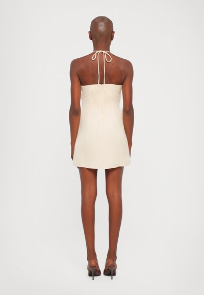 Strapless crèmekleurig mini-jurkje met een gesmokte achterkant en halterstrik. Het heeft een gladde textuur en een getailleerde silhouette, gecombineerd met hoge hakken.