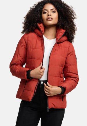 Vrouw met krullend haar die een rode gepofte jas met rits draagt over een wit shirt en zwarte broek tegen een effen achtergrond.