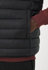 Chaleco acolchado negro con diseño de acolchado horizontal, que cuenta con un bolsillo lateral. Lleva puesto un camisa de manga larga roja con puños acanalados.