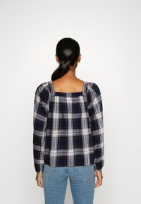 Chemisier à carreaux bleu marine, blanc et vert. Présente un décolleté carré et des manches bouffantes. Porté avec un jean bleu clair. Tissu en coton texturé.