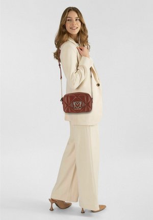 Bruine doorgestikte crossbodytas met een zilveren logo. Zacht leren materiaal, rechthoekige vorm en een kettingriem. Model draagt een licht pak.