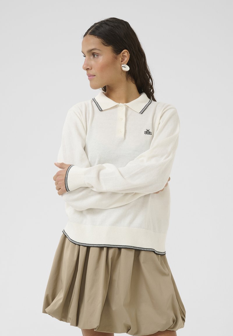 White long-sleeve polo shirt with black accents, featuring a buttoned collar and logo. Paired with a beige pleated skirt.