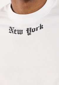 Camiseta blanca de algodón con un gráfico negro de "Nueva York" en una fuente audaz y ornamentada, situado cerca del escote.