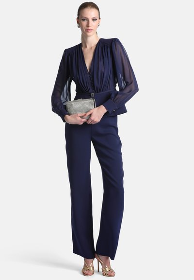 Femme en blouse bleu marine à manches transparentes et pantalon, tenant une pochette argentée, portant des talons hauts dorés à brides, face à l'avant.