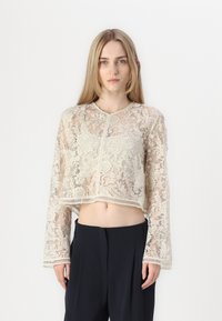 Haut en dentelle raccourci en blanc cassé avec des motifs floraux complexes, manches longues et col rond. Tissu texturé avec des bords ondulés.