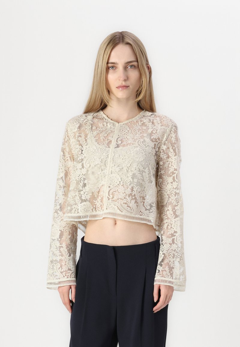 Haut en dentelle raccourci en blanc cassé avec des motifs floraux complexes, manches longues et col rond. Tissu texturé avec des bords ondulés.