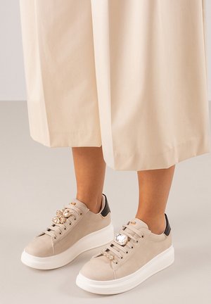 Zapatillas de ante beige con una suela blanca gruesa, adornadas con gemas decorativas en los cordones, con un talón negro en contraste.
