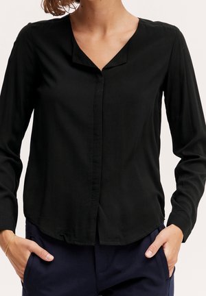 Frau trägt eine langärmelige schwarze Bluse mit V-Ausschnitt und dunkle Hose, Hände teilweise in den Taschen, vor einfachem Hintergrund.