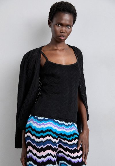 Black textured cardigan over a black tank top, paired with patterned multicolored zigzag pants in blue, pink, and black.
