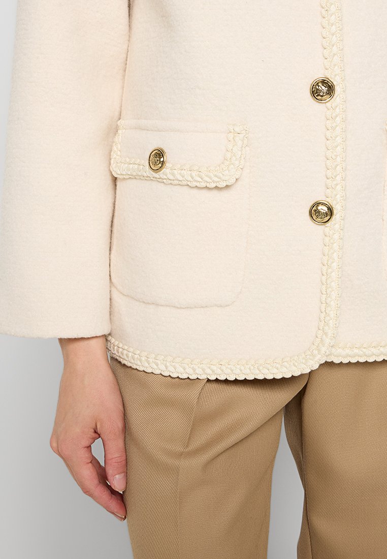 Cremfarbene Wollmischjacke mit strukturierter Oberfläche, goldfarbenen Knöpfen, geflochtenem Besatz entlang der Kanten und zwei vorderen Taschen.