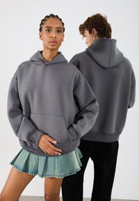 Sudadera con capucha gris y bolsillo frontal, presentada en un modelo. Tejido suave y texturizado con un corte holgado. Vista trasera mostrada con otro modelo.