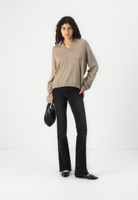 Pullover beige con scollo a V e maniche lunghe, abbinato a jeans a zampa nera. La modella tiene in mano una borsa nera e indossa ballerine nere con dettagli. 