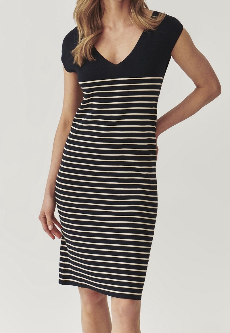 Femme portant une robe noire mi-longue à rayures horizontales beiges et manches courtes, debout avec une main sur la hanche.