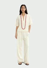 Donna in piedi che indossa un outfit cremoso e ampio con una maglietta a maniche corte, pantaloni larghi, sandali neri e una lunga collana di perline rosse.