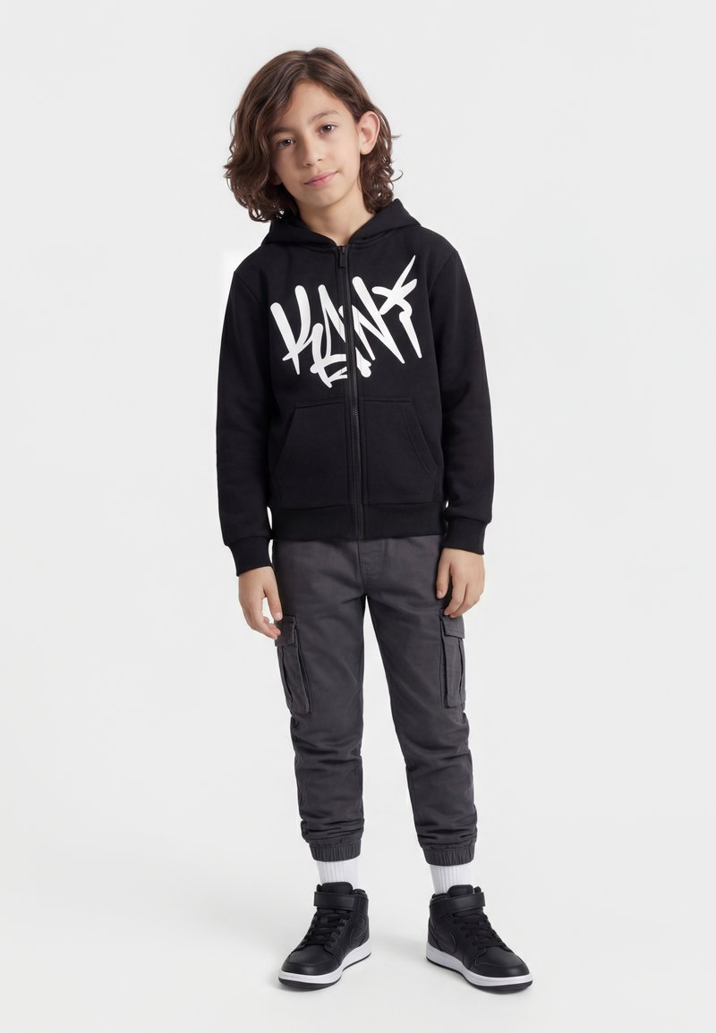 Giovane con capelli di media lunghezza che indossa una felpa con zip nera con stampa, pantaloni cargo grigi, calzini bianchi e sneaker alte nere in piedi.