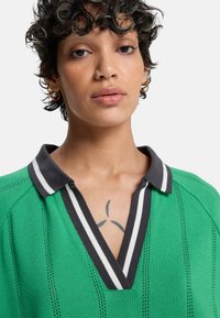 Polo vert en maille avec un col en V profond, un col rayé noir et blanc, et un motif texturé. Coupe ajustée avec des coutures visibles.