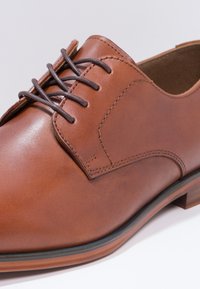 Chaussure en cuir marron avec une finition lisse, présentant un bout rond, un laçage à cinq œillets et des coutures contrastées.